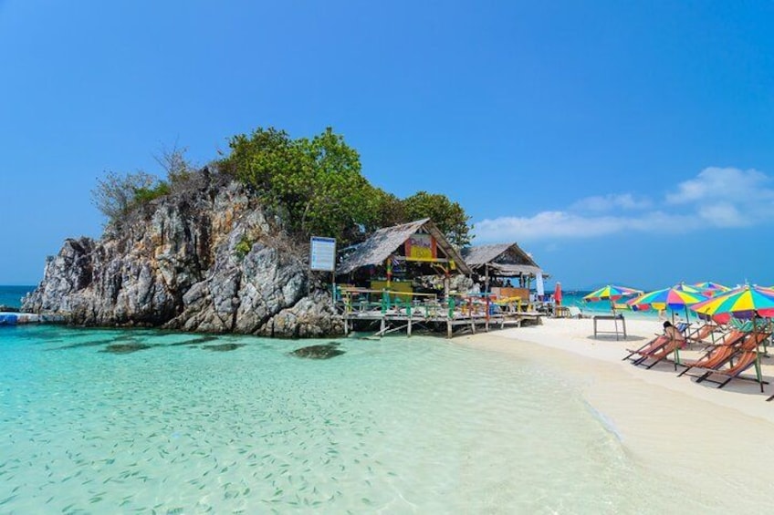 Een tropisch strand met helder turquoise water, een rotsformatie en een houten constructie met kleurrijke parasols. 4