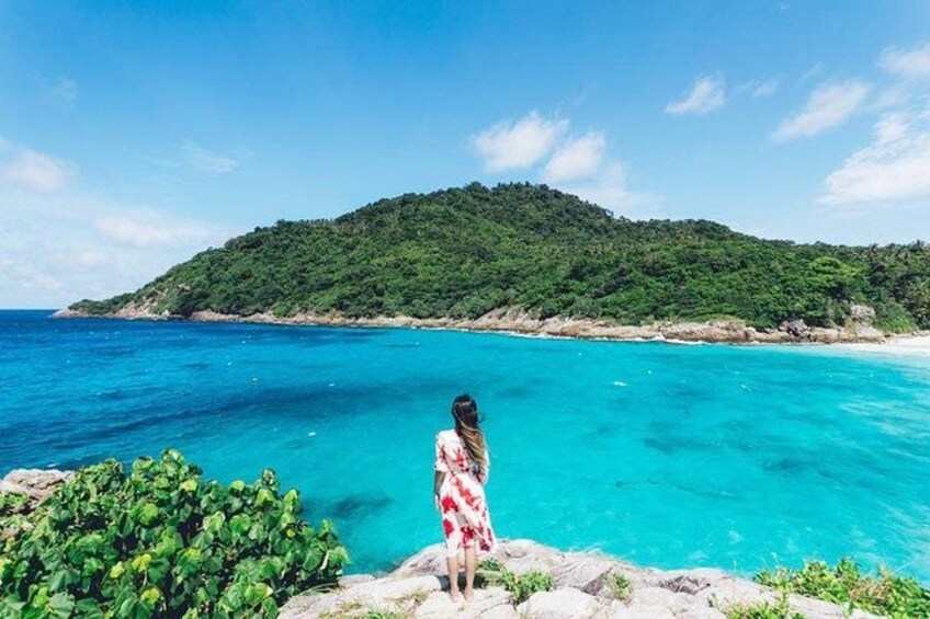 A woman in a red dress standing on a rocky outcrop overlooking turquoise waters and a green hill. 2