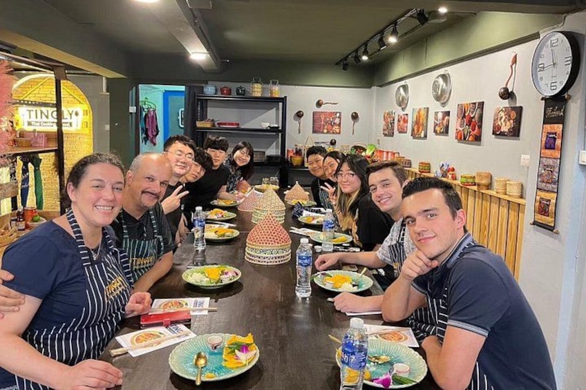 Un groupe de personnes est assis autour d’une table, devant de la nourriture et des boissons, dans une pièce où l’on peut voir des étagères et des décorations murales. 1