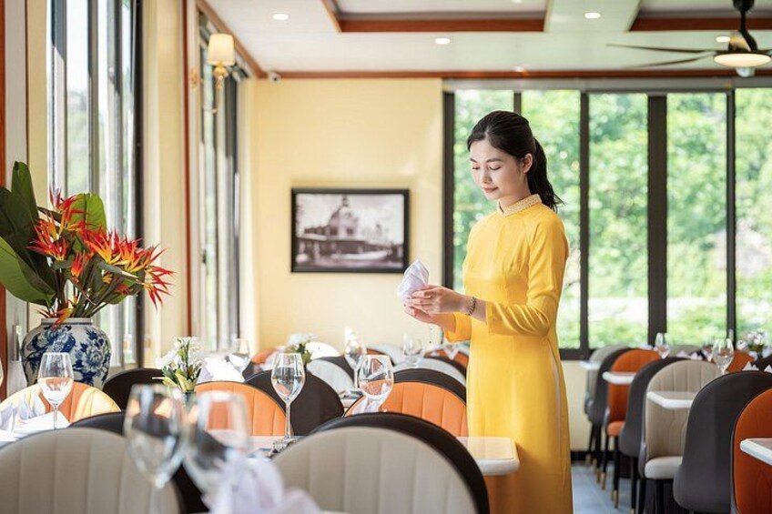 A person in a yellow dress standing in a restaurant with tables set for dining, a vase with flowers, and a ceiling fan. 3