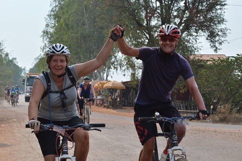 Two cyclists on a dirt road with trees and a building in the background. 1