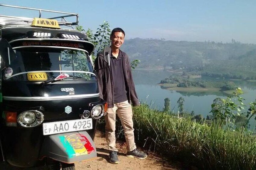 A man standing next to a black auto-rickshaw with a license plate, a scenic view of a lake and hills in the background. 2