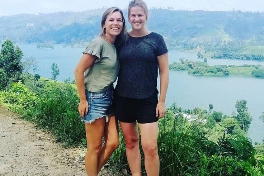 Two people standing on a path with a lake and hills in the background. 1