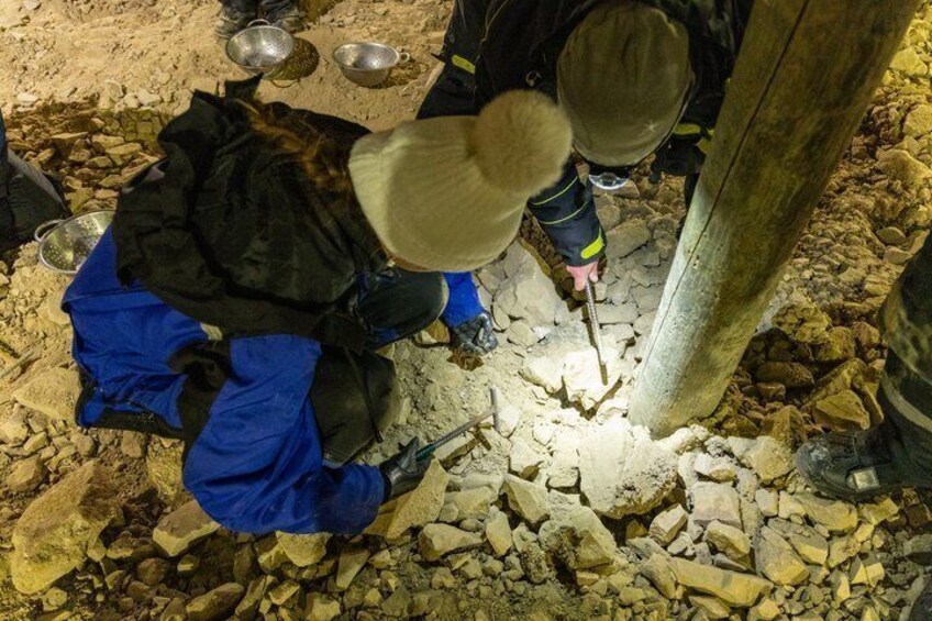 Two people digging in a rocky area with tools and metal bowls. 3