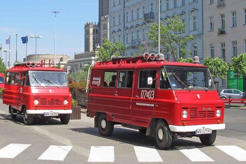 Two red vintage vans parked on a street with buildings and trees in the background. 1