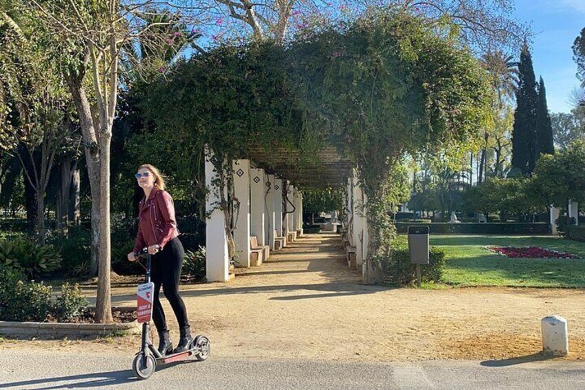 A person riding a scooter on a paved path in a park with trees and a pergola. 2