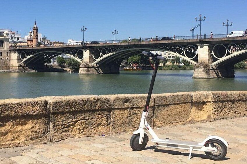 A white electric scooter parked on a stone wall by a river, with a historic bridge in the background. 4