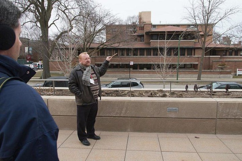 A man gesturing while speaking to a group of people outdoors, with buildings and a parking lot in the background. 4