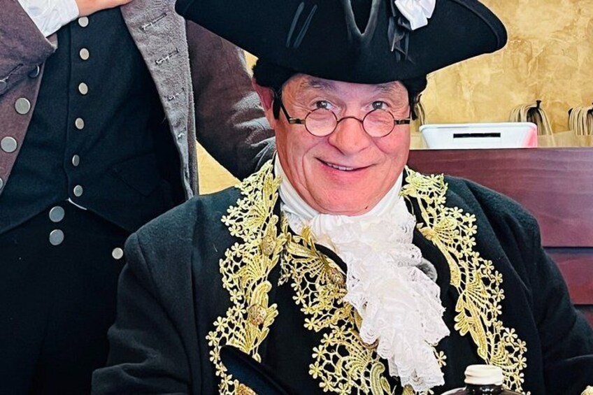 A person in historical attire with a black coat, white cravat, and a black hat with a white plume. 4