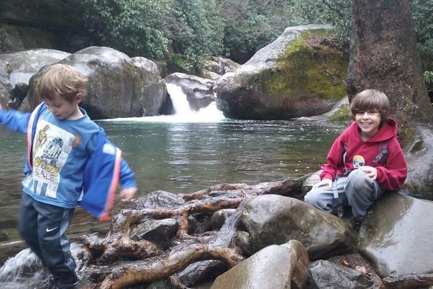 Two children playing near a waterfall in a forested area with large rocks and a flowing stream. 1