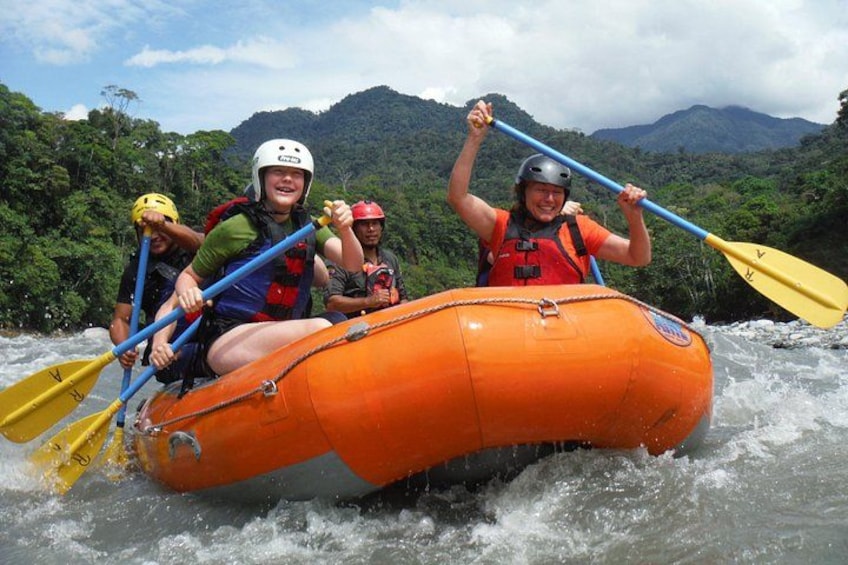 A group of people rafting on an orange raft with a river and mountains in the background. 1