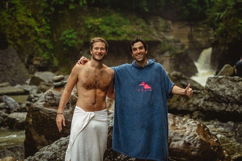 Two people standing on rocks near a waterfall in a forested area. 4