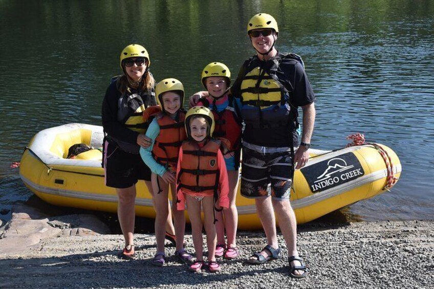 A group of people wearing helmets and life jackets standing by a yellow inflatable raft on a gravel shore by a body of water. 2