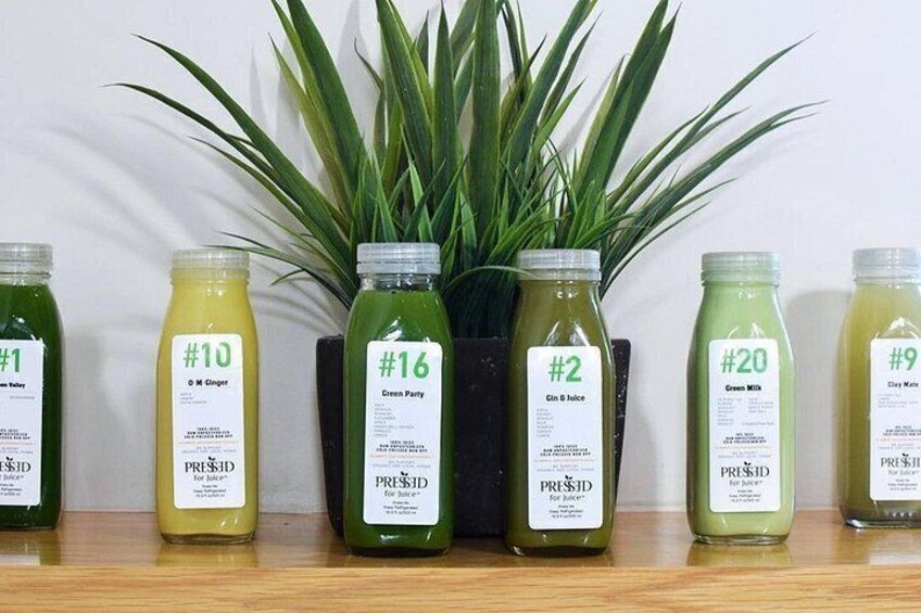Six bottles of juice with labels, placed on a wooden shelf with a plant in the background. 3