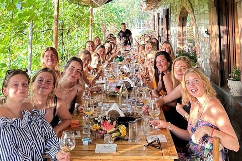 A group of people seated at a long wooden table outdoors, enjoying a meal with wine glasses and various dishes. 5