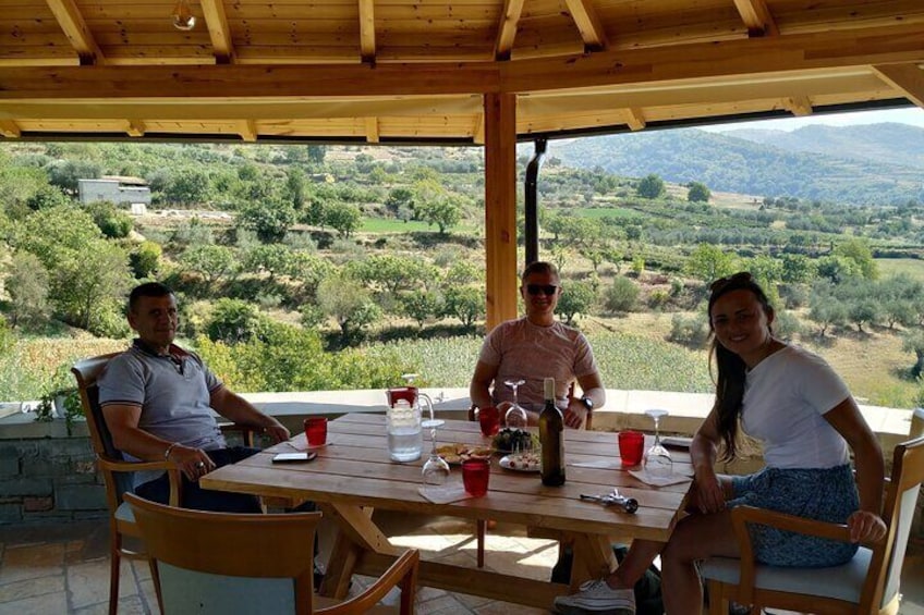 Three people seated at an outdoor wooden table with a scenic view of a hilly landscape in the background. 4