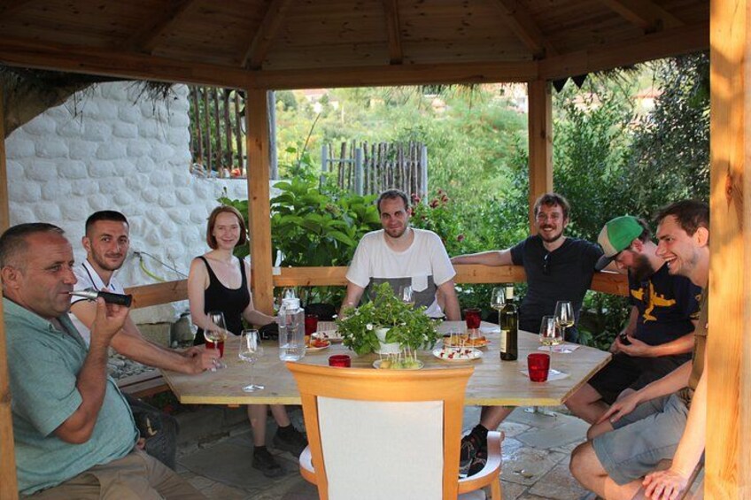 A group of people sitting at an outdoor wooden table with food and drinks, surrounded by greenery. 5