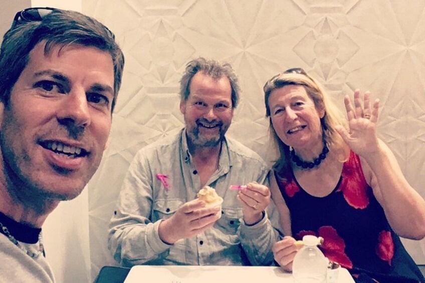 Tres personas sentadas a una mesa con comida, una de ellas sosteniendo una bebida, y una pared con diseño en el fondo. 2