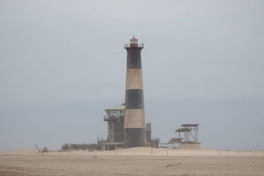 A tall lighthouse with a black and white striped tower, a control building, and a sandy beach in the background. 1