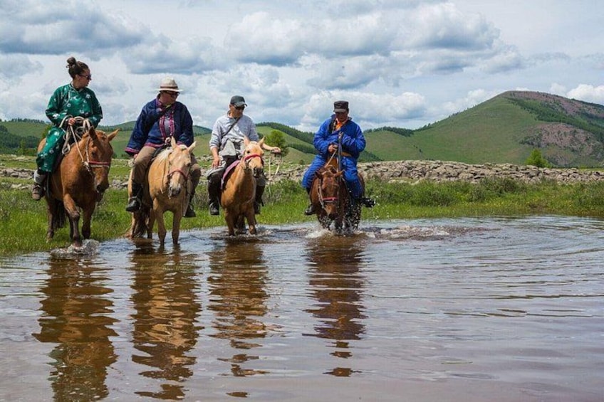 Three people riding horses through a shallow river in a grassy landscape with hills in the background. 2