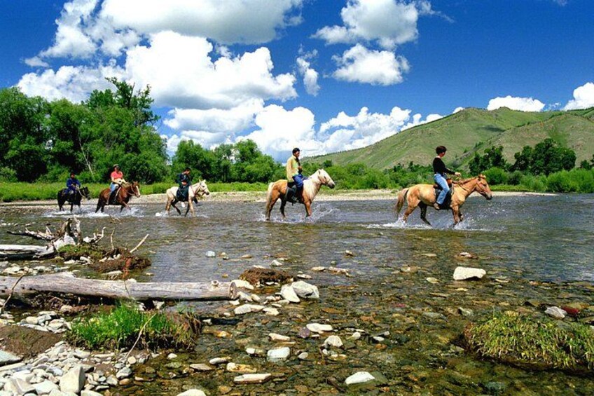 People riding horses across a shallow river with rocks and greenery in the background. 1