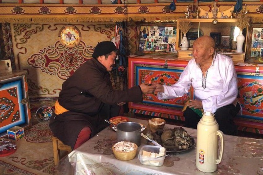 Two men sitting at a table with various dishes and a large jug, in a room with ornate decorations and a patterned rug. 5