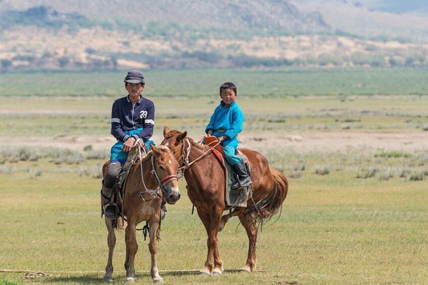 Two people riding horses in a grassy field with mountains in the background. 2