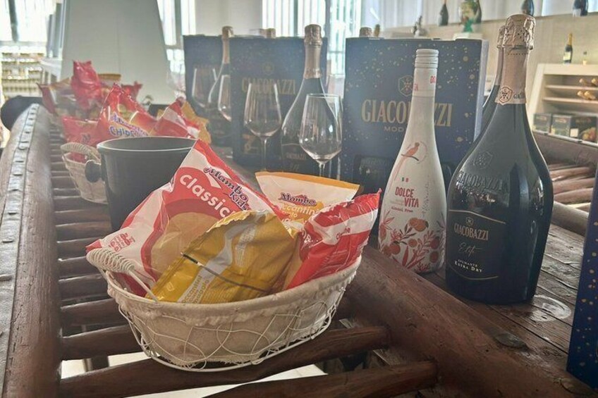 A wooden table with a basket of snacks, a bottle of Dolce Vita wine, and a bottle of Acqualazzoli wine. 3