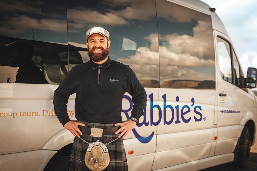 A man in a black shirt and kilt standing in front of a white van with 'Rabbie's' written on it. 4