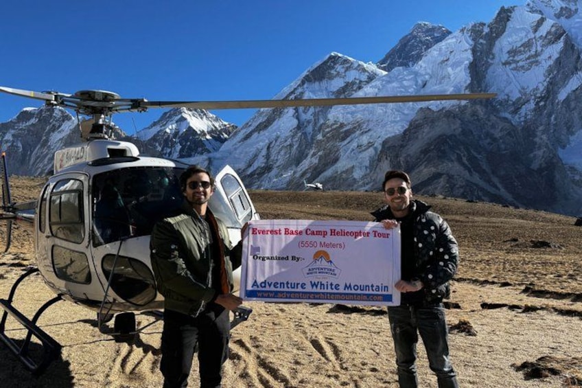 Two people holding a sign in front of a helicopter with snow-capped mountains in the background. 1