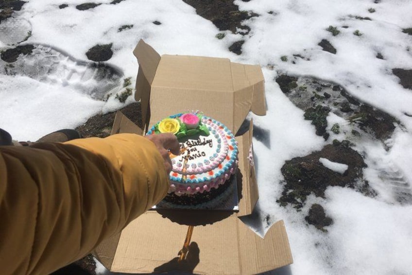 A hand holding a birthday cake in a cardboard box on snow-covered ground. 2