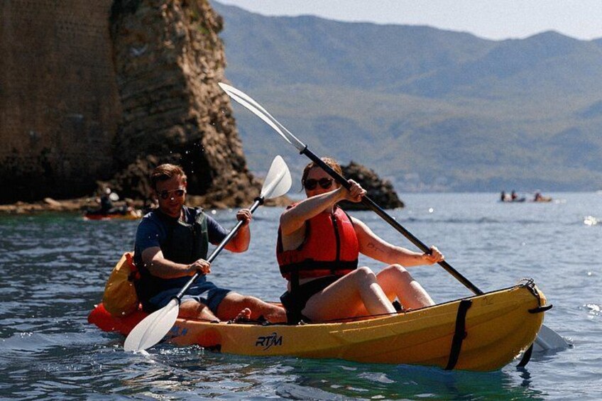 Budva Bay Kayak & SUP Tour to Coastal Caves – couple paddling an orange tandem kayak with mountain backdrop in Budva Bay 2