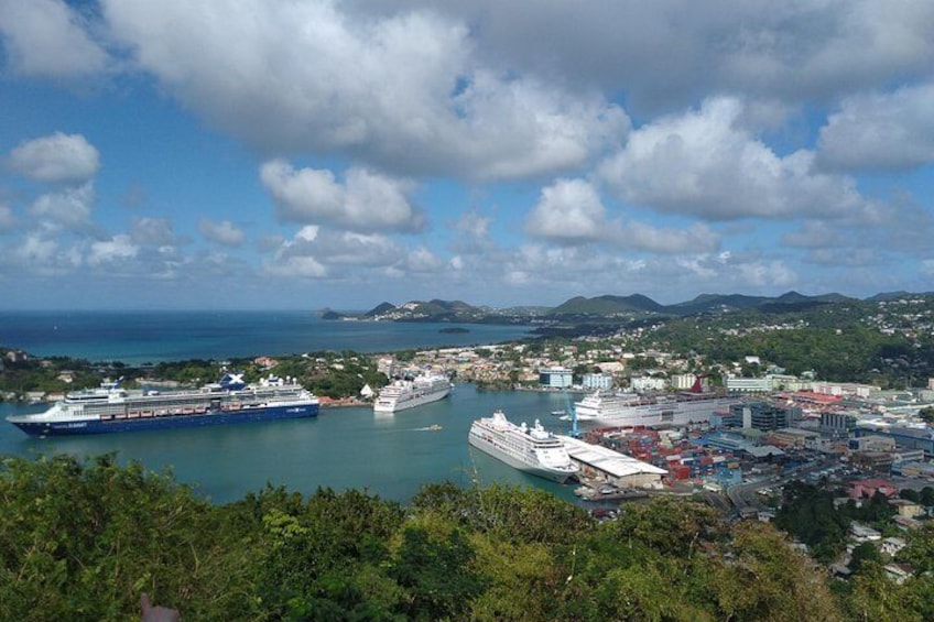 A coastal city with two large cruise ships docked at a port, surrounded by lush greenery and hills in the background. 2
