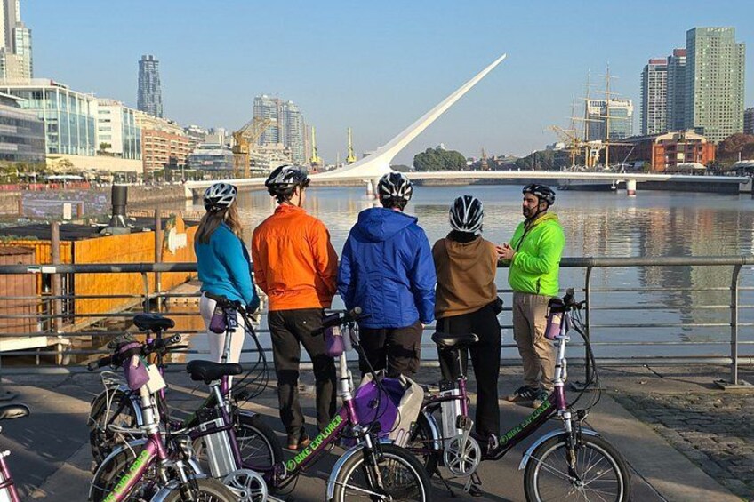 A group of cyclists standing by a waterfront with a modern bridge in the background. 5