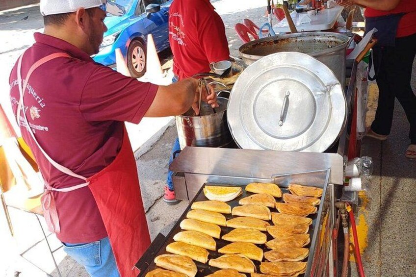 Un vendeur de nourriture de rue prépare des plats dans un étal, avec une grande marmite en métal et un plateau rempli d’aliments cuits. 2