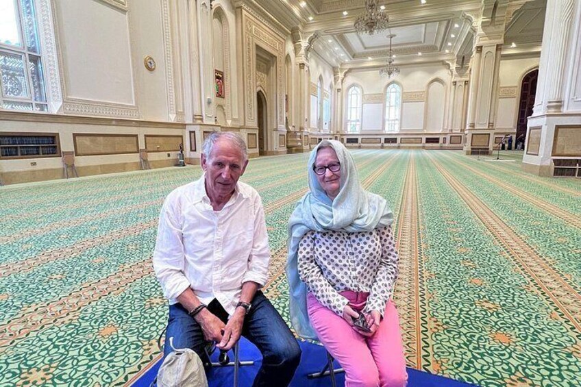 Two people sitting on a carpeted floor in a large hall with patterned flooring and arched windows. 4