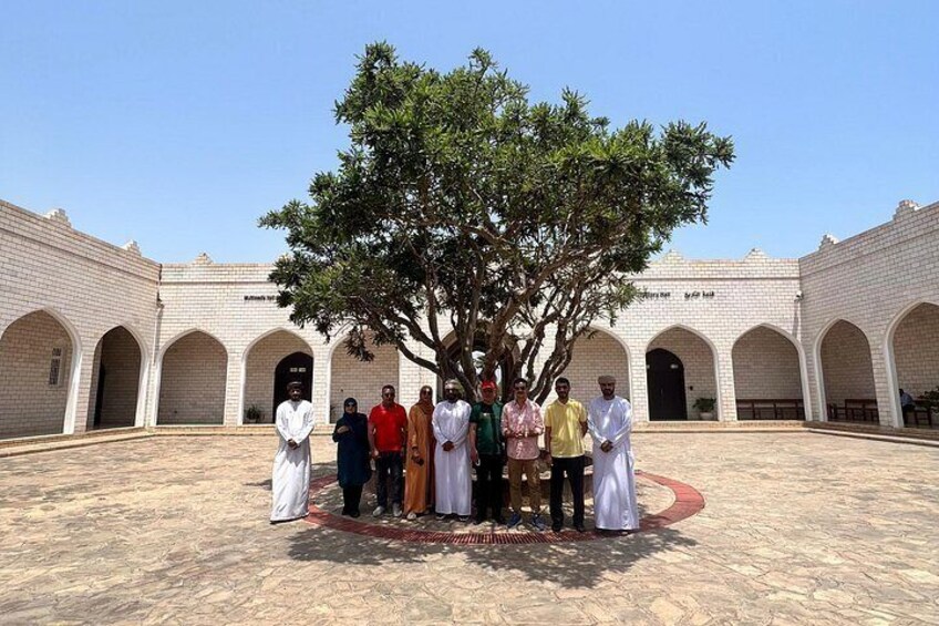 A group of people standing under a tree in a courtyard with a white building in the background. 5