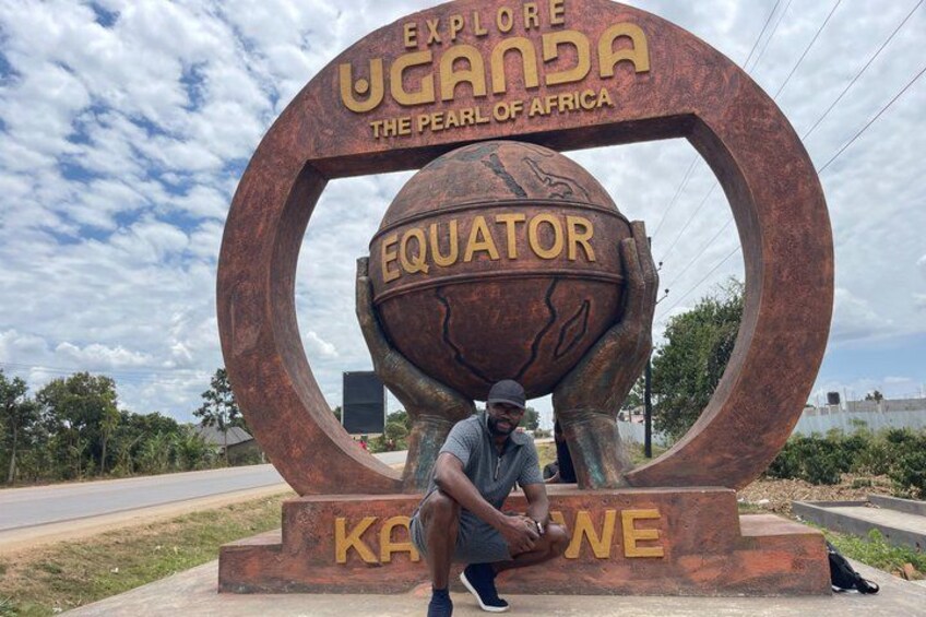 Una grande scultura in metallo con il testo 'EXPLORE UGANDA THE PEARL OF AFRICA' e 'EQUATOR' al centro, e una persona seduta di fronte. 1