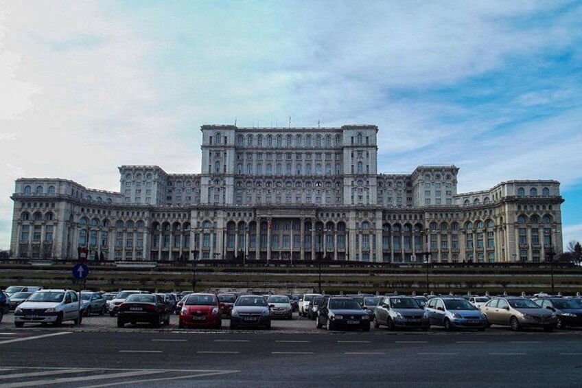 A large, historic building with multiple levels and arched windows, surrounded by a parking lot with various cars. 4