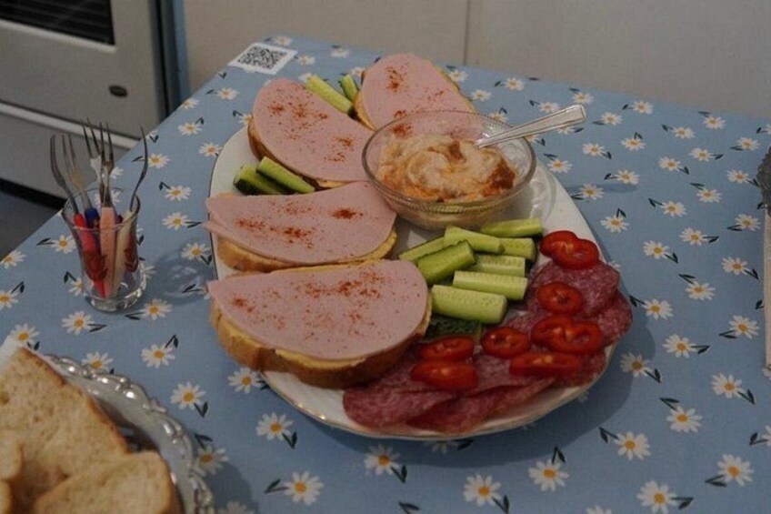 A table with a floral tablecloth, a plate of sliced ham, cucumber sticks, cherry tomatoes, and a bowl of dip. 5