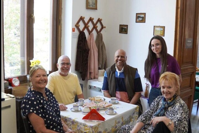A group of people seated around a table with food and drinks, in a room with a window and a coat rack. 1