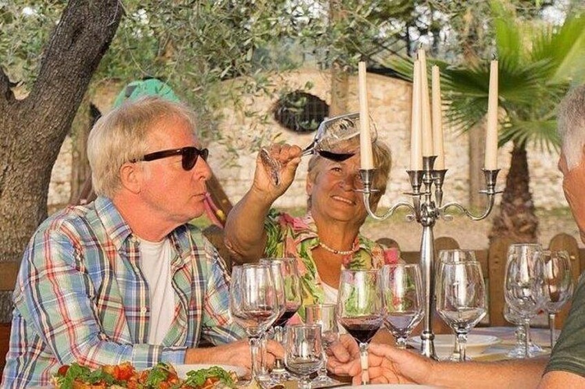 A group of people dining outdoors with a table set for a meal, surrounded by greenery and a candelabra. 3