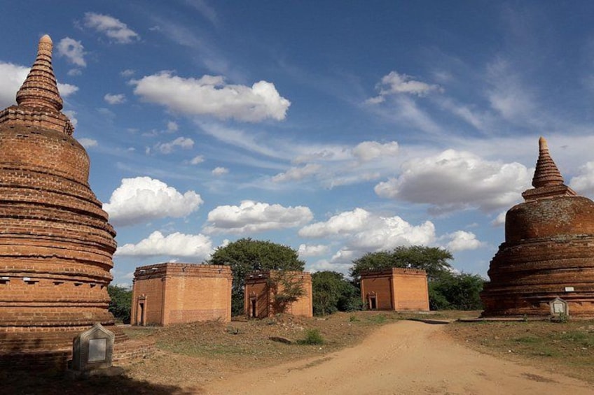 A dirt path leading to ancient brick structures with conical roofs, surrounded by trees and a clear sky. 2
