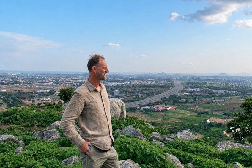 A man standing on a rocky outcrop overlooking a cityscape with a river and hills in the distance. 2