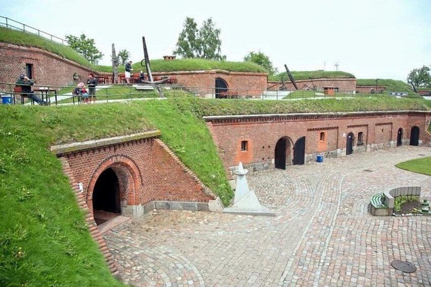 En historisk fæstning med murstensmure, grønne græsdike og en belagt gårdsplads. 2