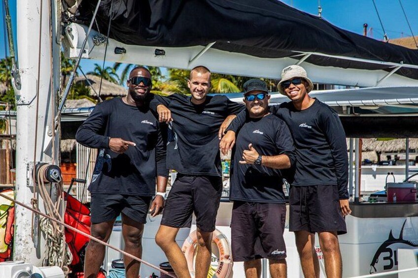 Four people on a boat, wearing matching black outfits, with a clear sky and palm trees in the background. 2