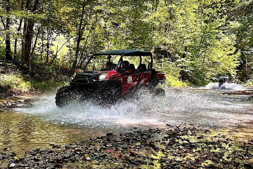 A red off-road vehicle driving through a shallow stream in a forested area. 2
