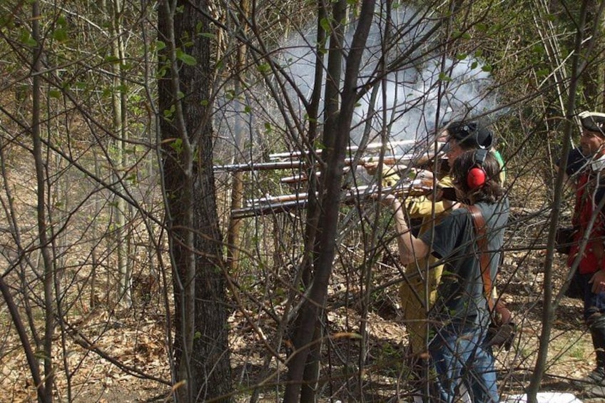 A person aiming a rifle in a wooded area with trees and a clear sky. 2