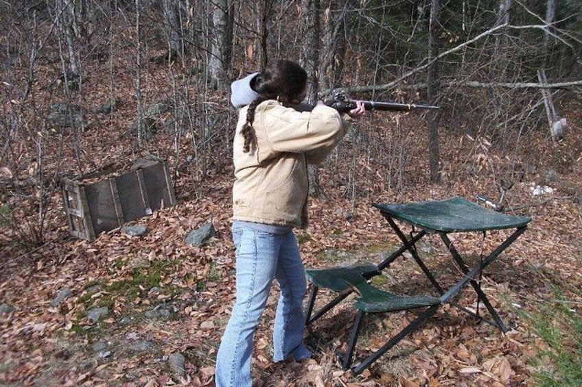 A person aiming a rifle in a wooded area with a folding chair and a box in the background. 4