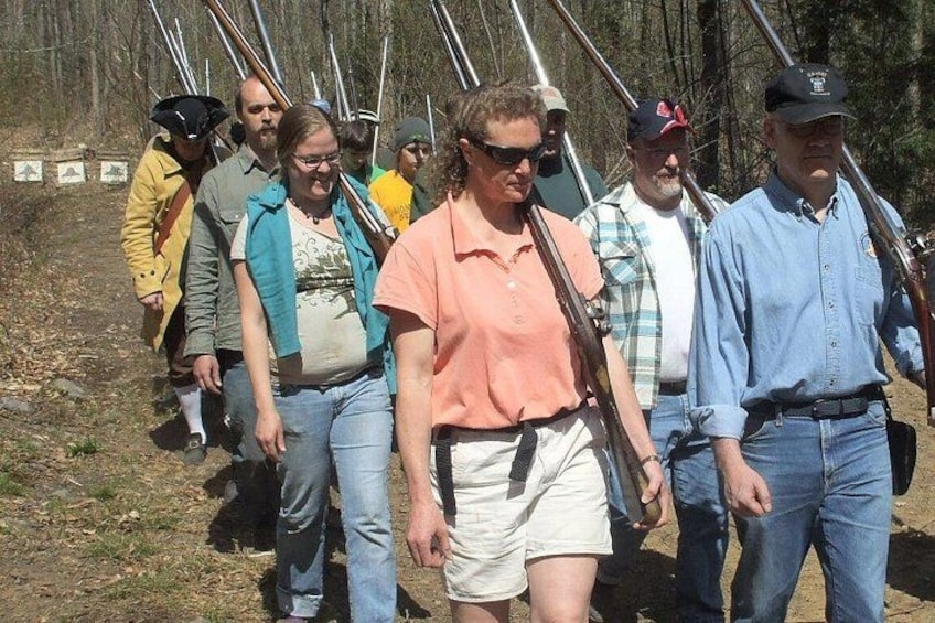 A group of people walking outdoors, some carrying rifles, in a wooded area. 1